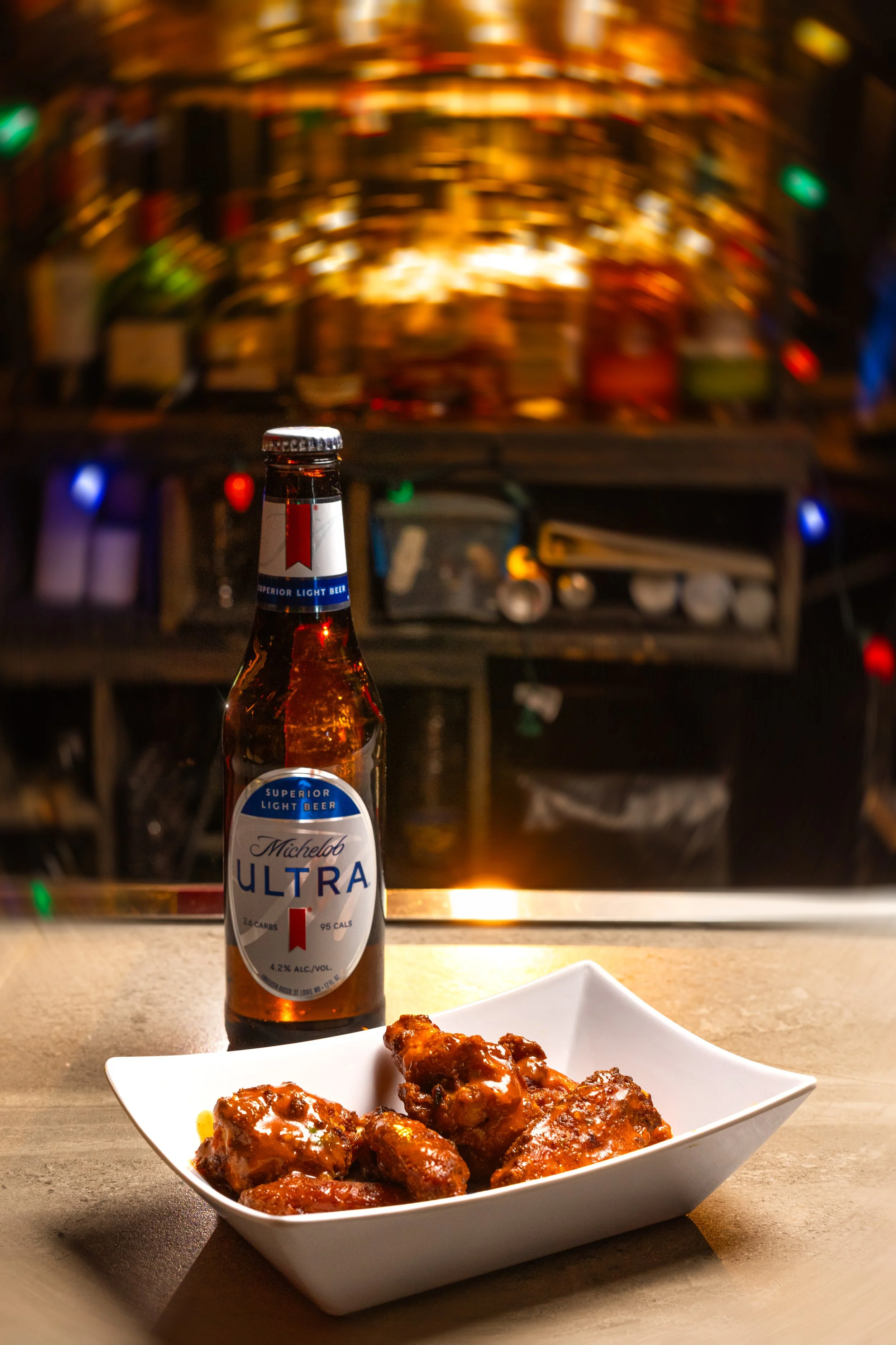 A bottle of Michelob Ultra beer next to a white bowl of saucy chicken wings on a bar counter, with a blurred background of colorful lights.