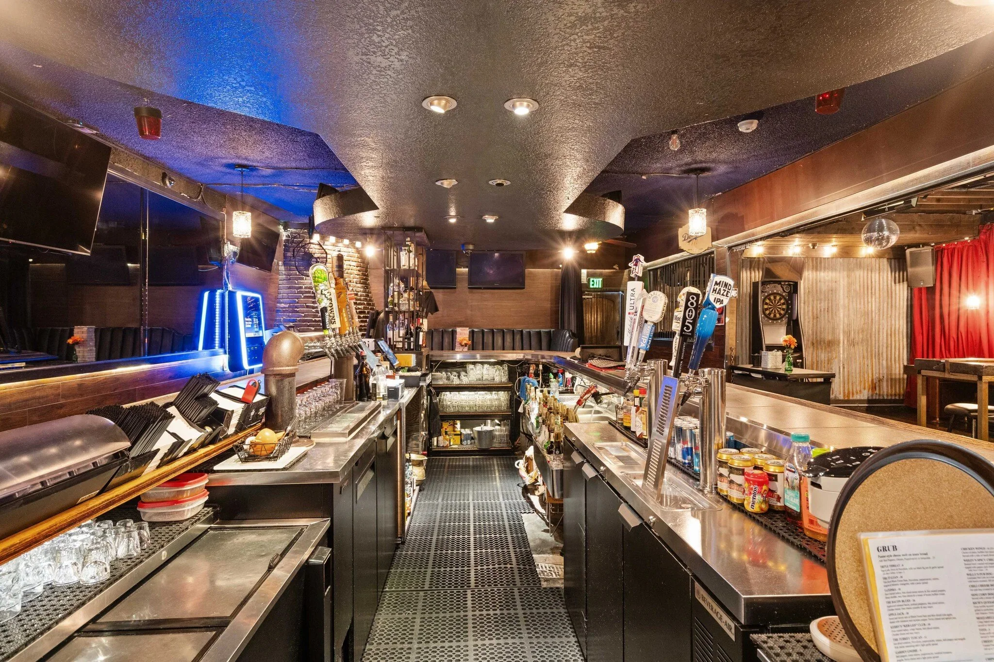 Empty bar with various beer taps, liquor bottles, and glasses; lounge with tables, red curtains, and dartboard in the background.