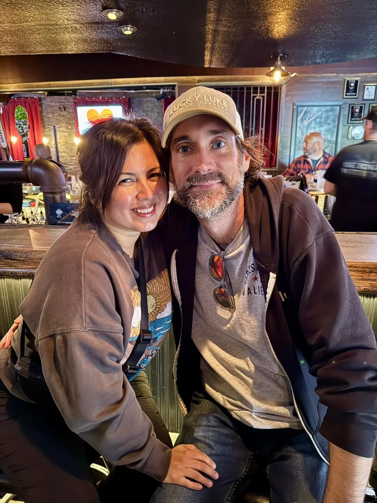 A woman and a man posing together and smiling inside a bar or restaurant, with people and a television in the background.
