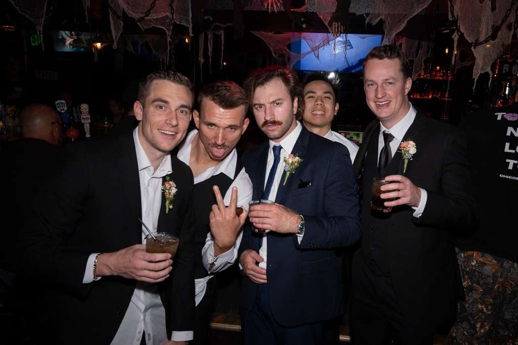 Group of five men in suits at a party or bar, some holding drinks, celebrating, with Halloween decorations like spider webs hanging overhead.