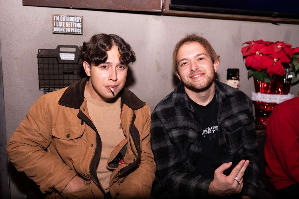 Two young men sitting indoors, with one smoking a cigarette. The man on the left wears a tan jacket and the man on the right wears a black and gray checkered shirt. There is a red poinsettia plant and a Santa decoration in the background.
