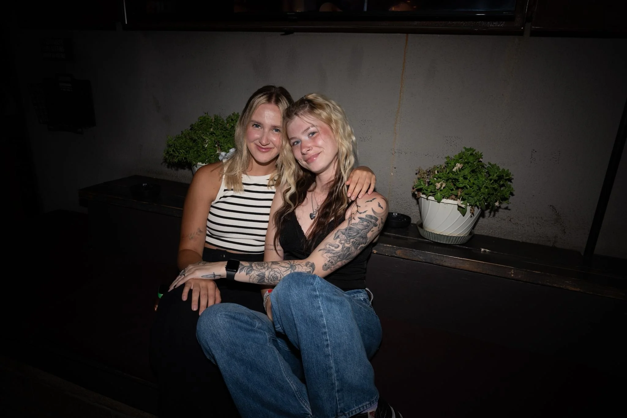 Two young women sitting together, one with blonde hair wearing a striped top and the other with wavy hair and tattoos, smiling and embracing each other, in a dimly lit indoor setting with plants in the background.
