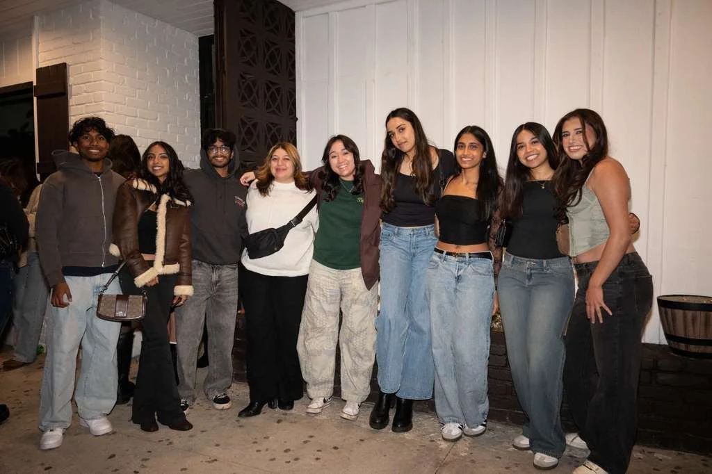 Group of nine young adults standing together indoors, smiling, with a white paneled wall behind them.