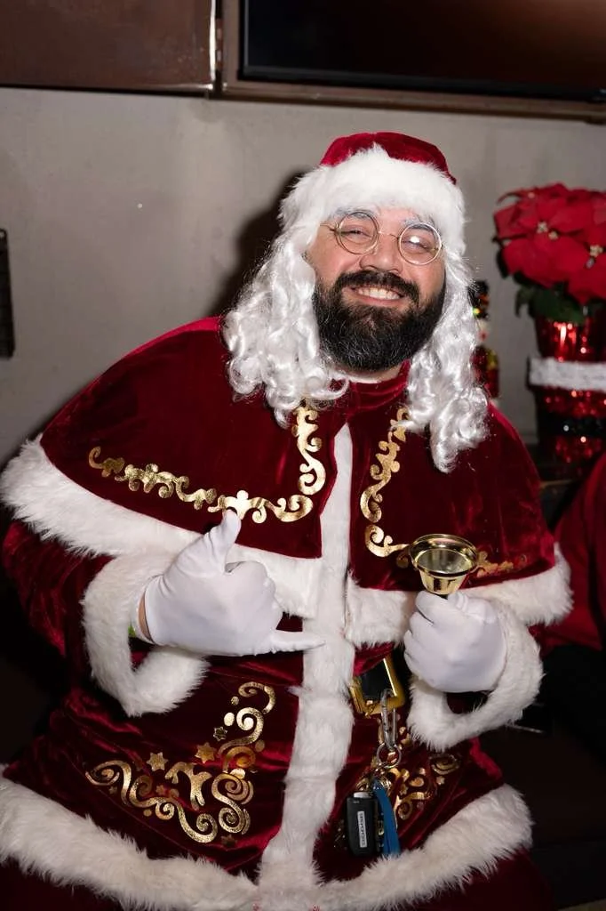 A man dressed as Santa Claus with a white beard, round glasses, and a red and white costume, smiling and pointing at himself while holding a small gold bell, in a festive setting with poinsettia decorations.