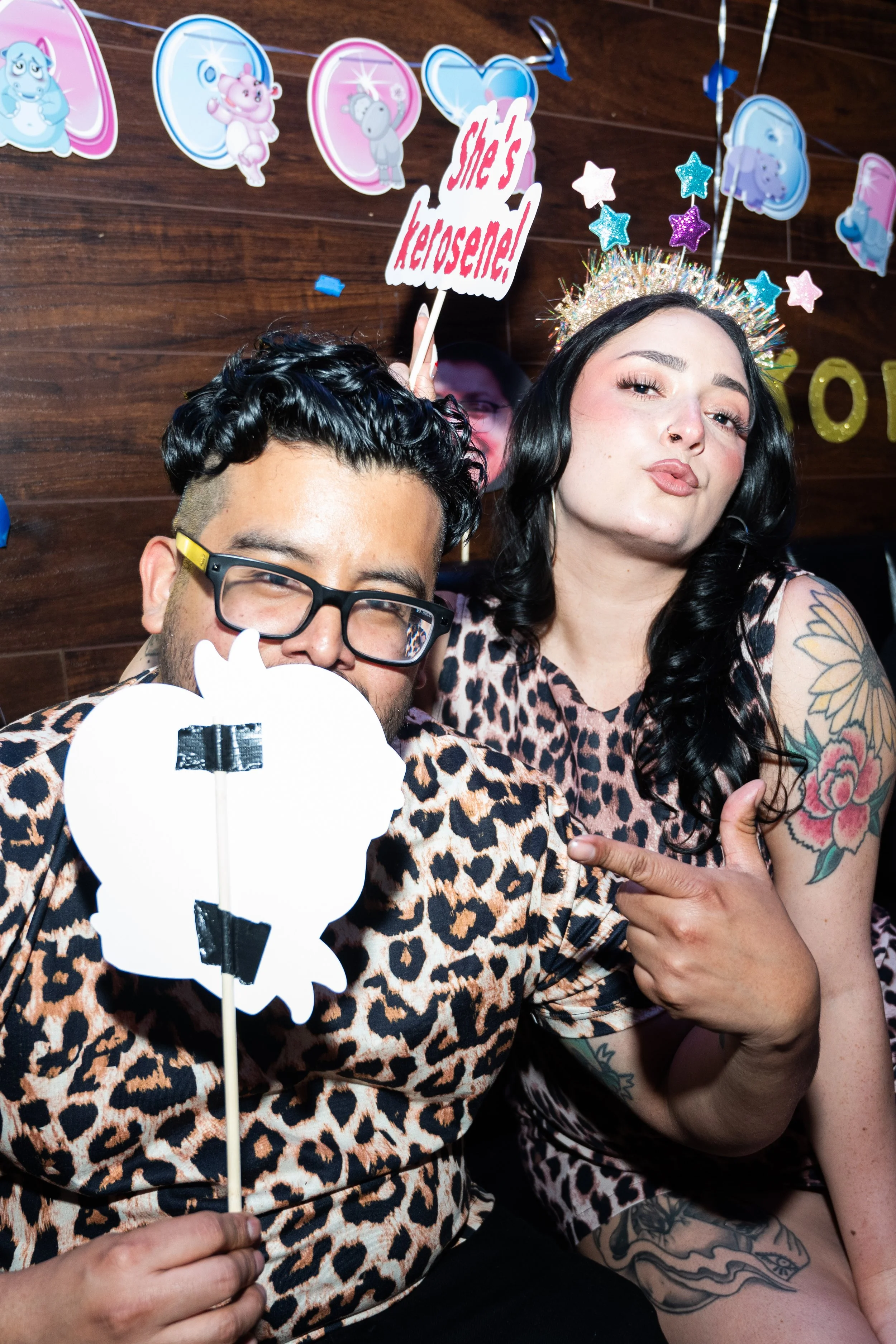 Two people in leopard print clothing at a party with playful expressions, one holding a sign saying "She's kerosene!" Backdrop of cartoon heart decorations.