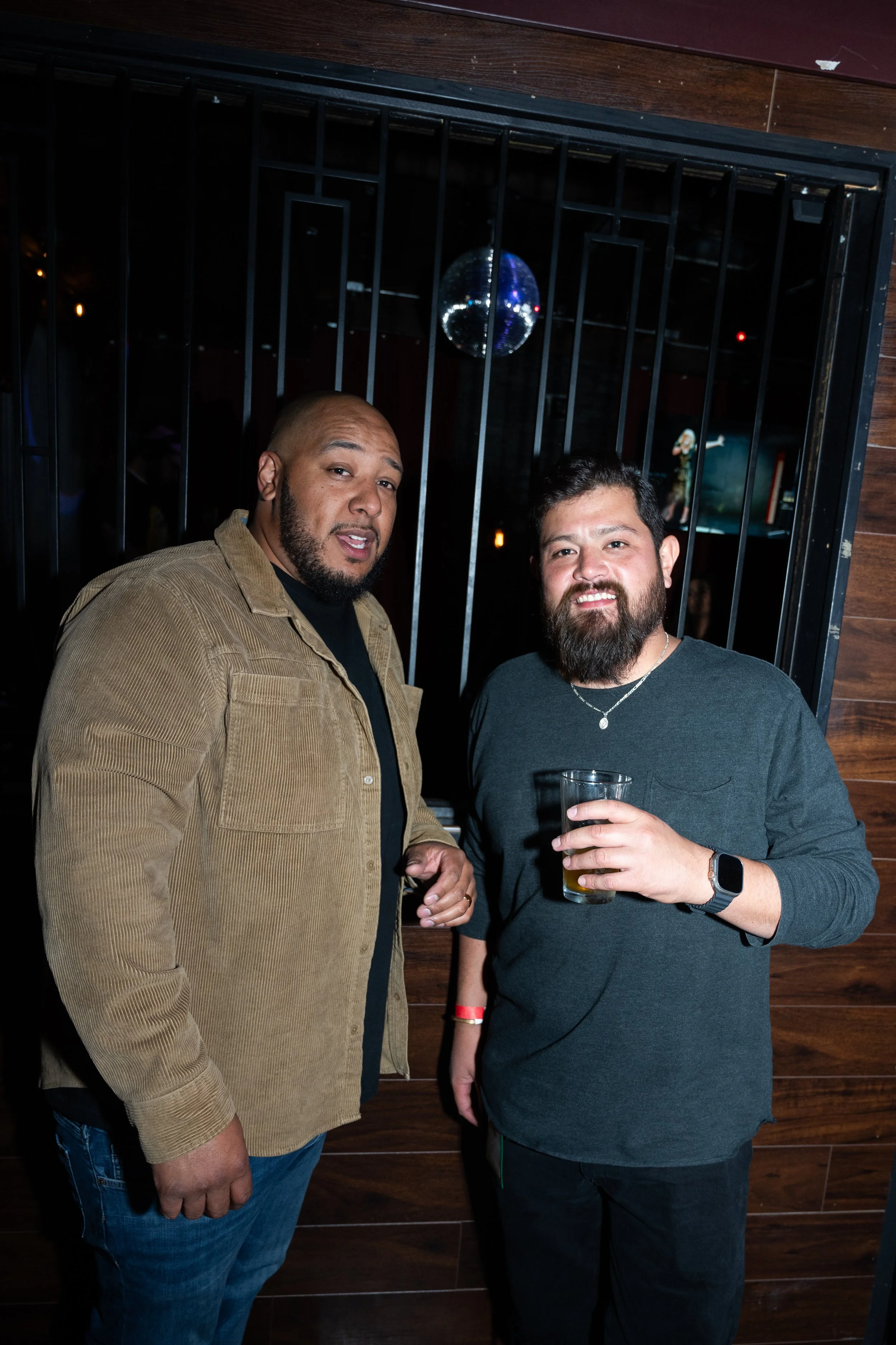 Two men standing near a wooden wall, one holding a drink, in a dimly lit room with a disco ball overhead.