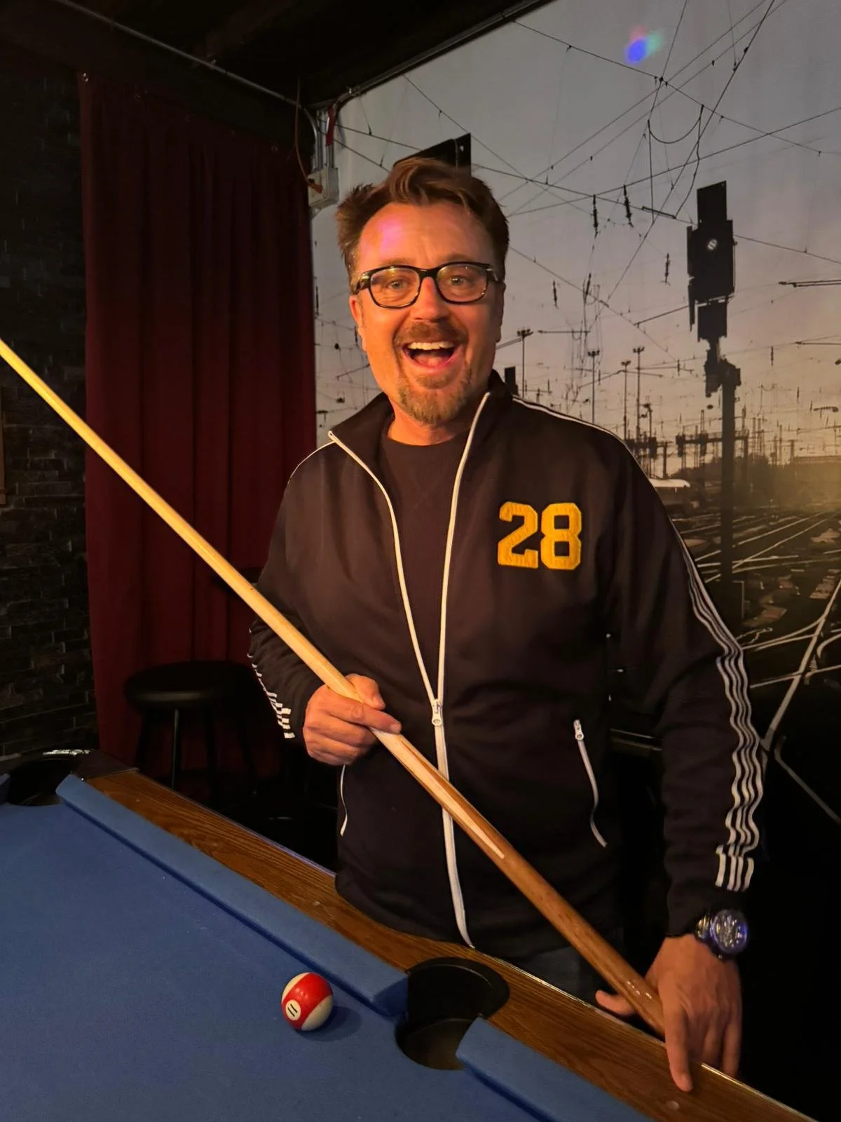 A man with glasses, a beard, and a mustache smiling cheerfully, holding a pool cue, standing by a billiards table in a dimly lit room.