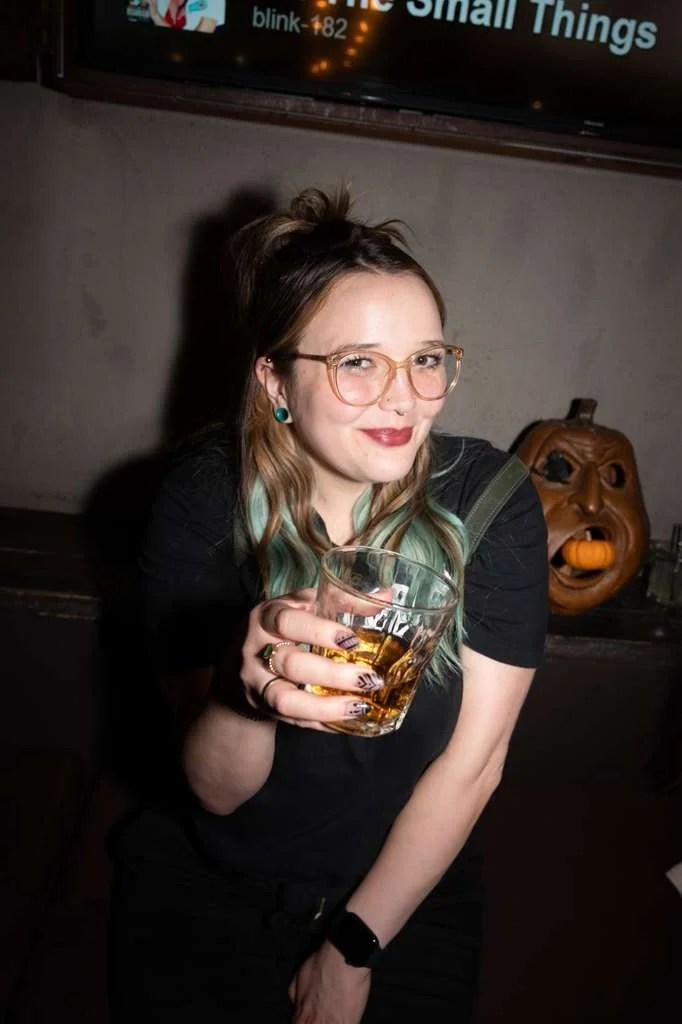 A young woman with glasses, styled hair, and dark lipstick holding a glass of drink, smiling at the camera, with a pumpkin decoration in the background.