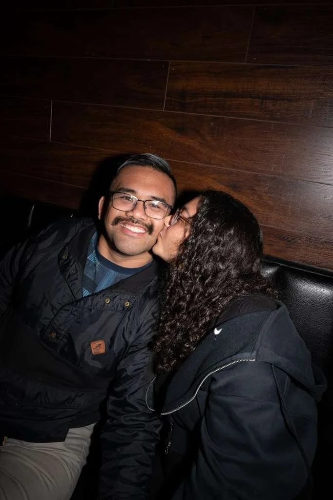 A woman is kissing a man on the cheek in a cozy indoor setting with wooden wall paneling in the background.
