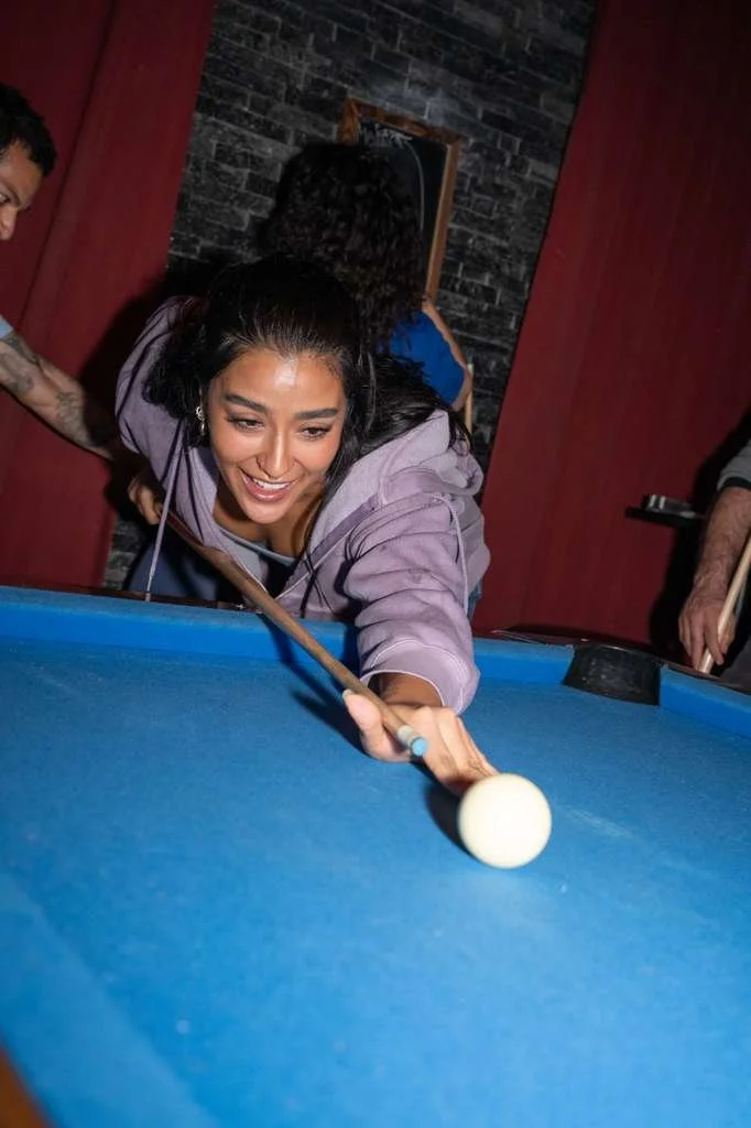 Woman smiling and aiming a cue stick to hit a white cue ball on a blue pool table.