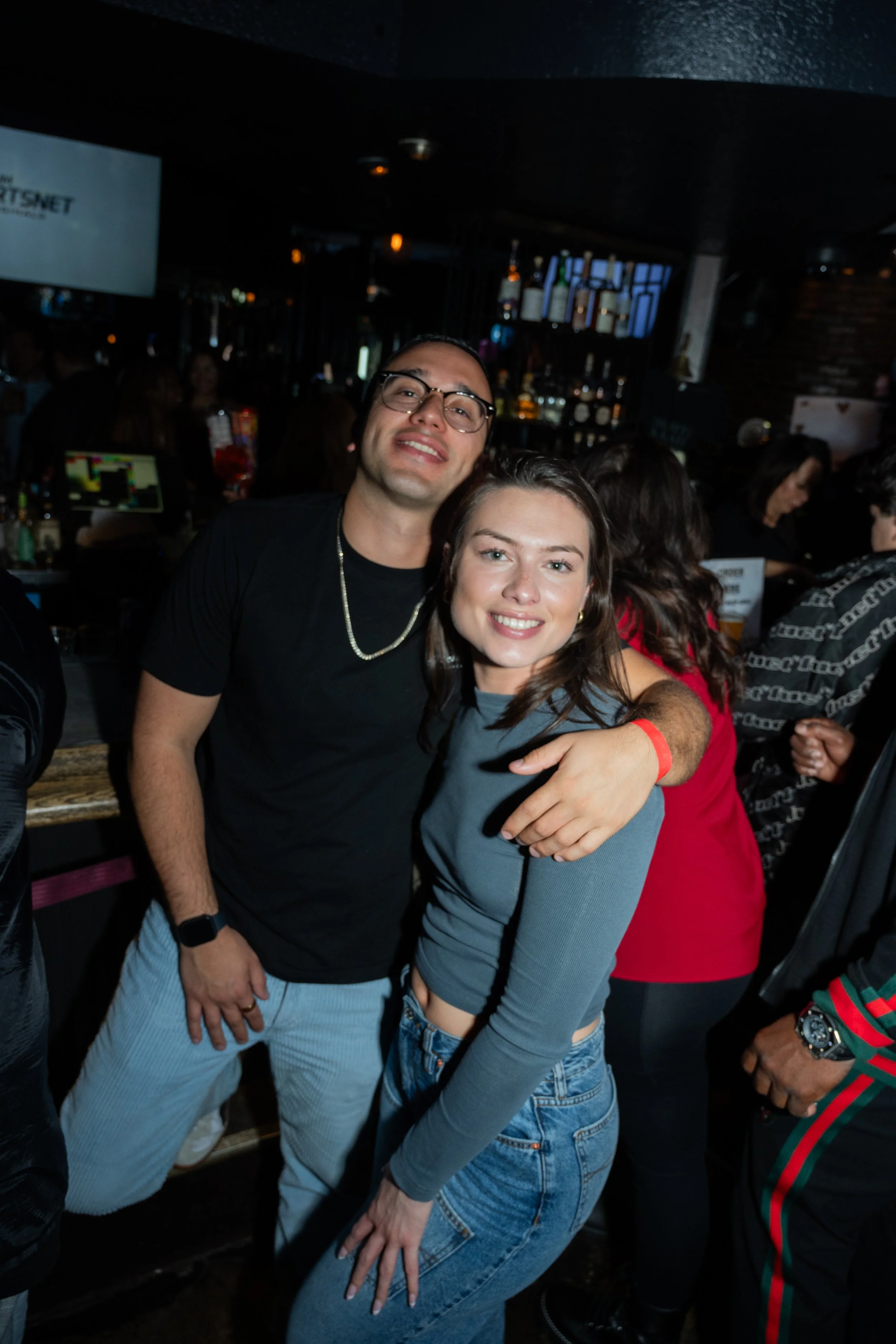 A man and woman posing together in a bar setting, with other people in the background. The man is wearing glasses and a black outfit, while the woman is in a long-sleeve gray top and jeans.