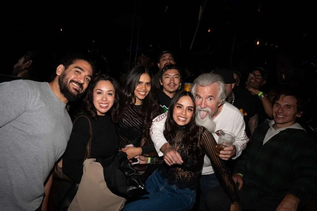 Group of smiling people at a social gathering, some holding drinks, in a dimly lit environment.