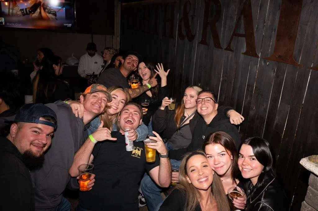 Group of young adults enjoying drinks at a bar or pub, smiling and posing for the photo in a dimly lit, rustic setting.