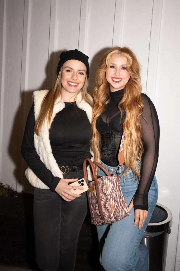 Two young women standing side by side indoors, smiling at the camera with a plain wall background. One has long blonde hair, and the other has long red hair. They are dressed stylishly, with one wearing a black beanie, black top, and white vest, hold