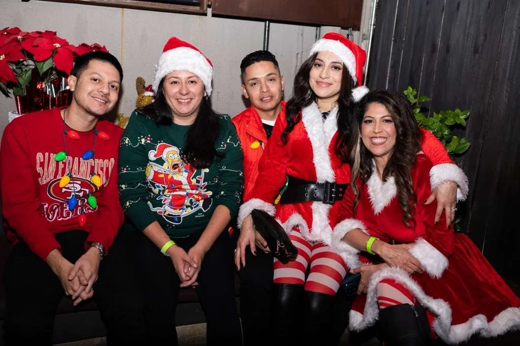 Group of five people dressed in Christmas-themed clothing, sitting together and smiling, with Christmas decorations and poinsettias in the background.