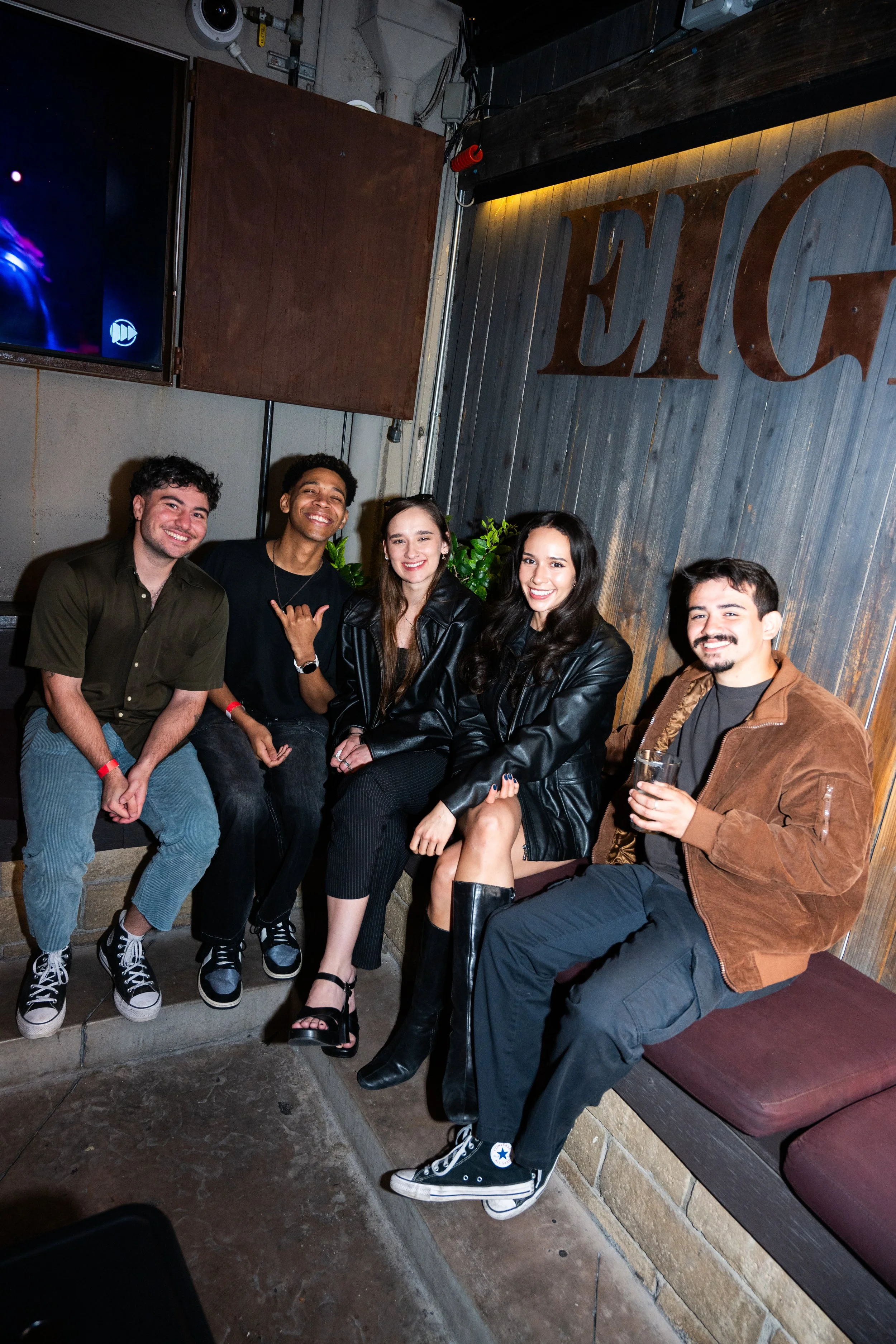 A group of five friends sitting and posing for a photo in a casual indoor setting, some wearing dark jackets and casual clothing.