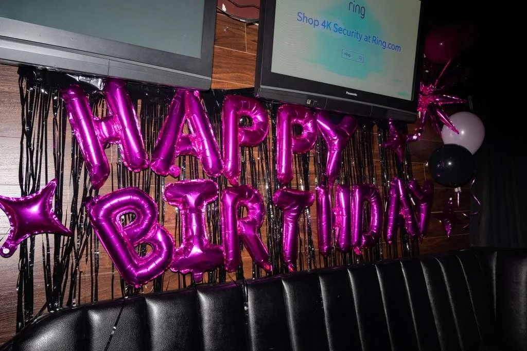 Pink metallic balloons spelling out 'HAPPY BIRTHDAY' with black streamers and additional balloons in black, white, and pink, in a decorated party setting.