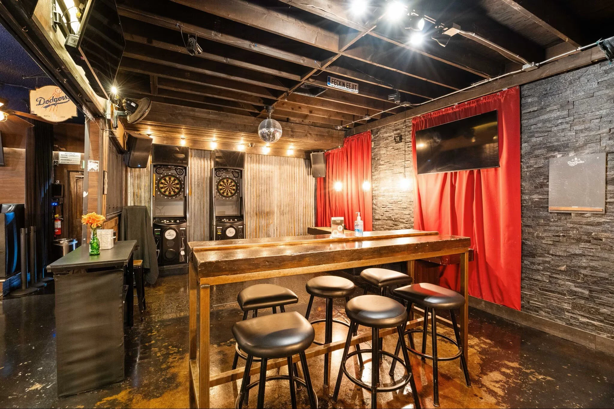 Bar area with dartboards, high table with bar stools, red curtains, TV, and disco ball in a dimly lit setting.