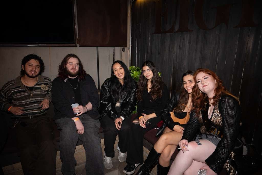 Six young adults sitting closely together on a bench in a dimly lit room, smiling and posing for the camera.