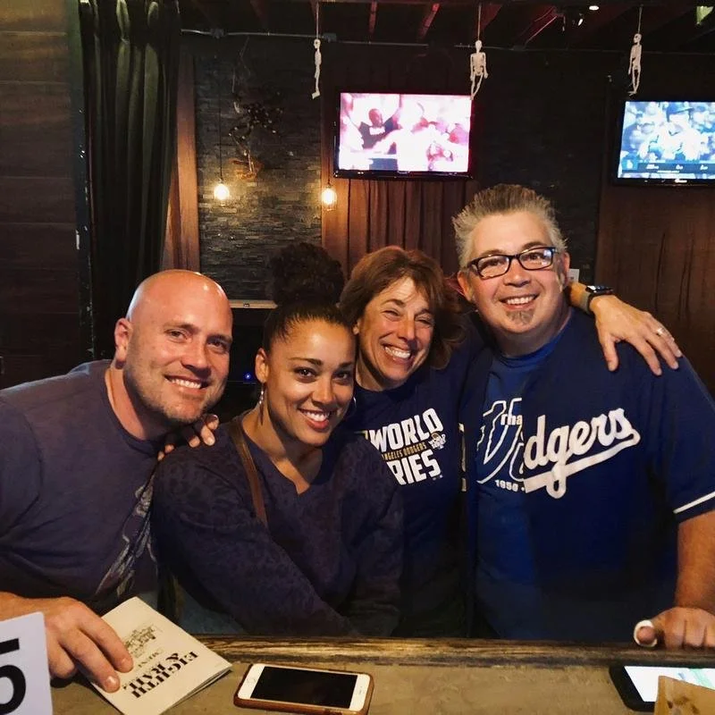 Four people smiling in a bar with sports game on TV in background.