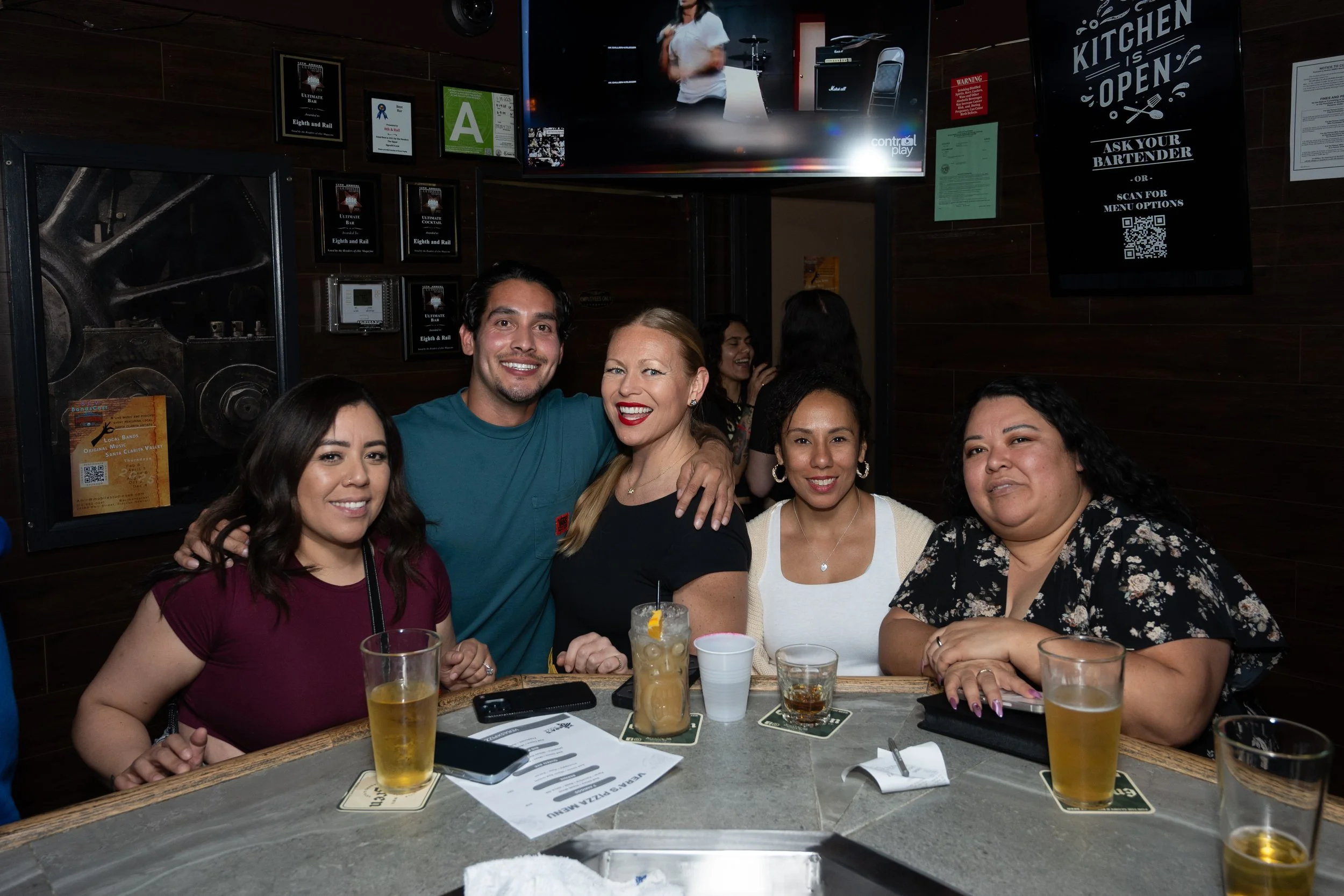 Group of people smiling at a bar, drinks on the counter, TV and framed certificates on the wall.
