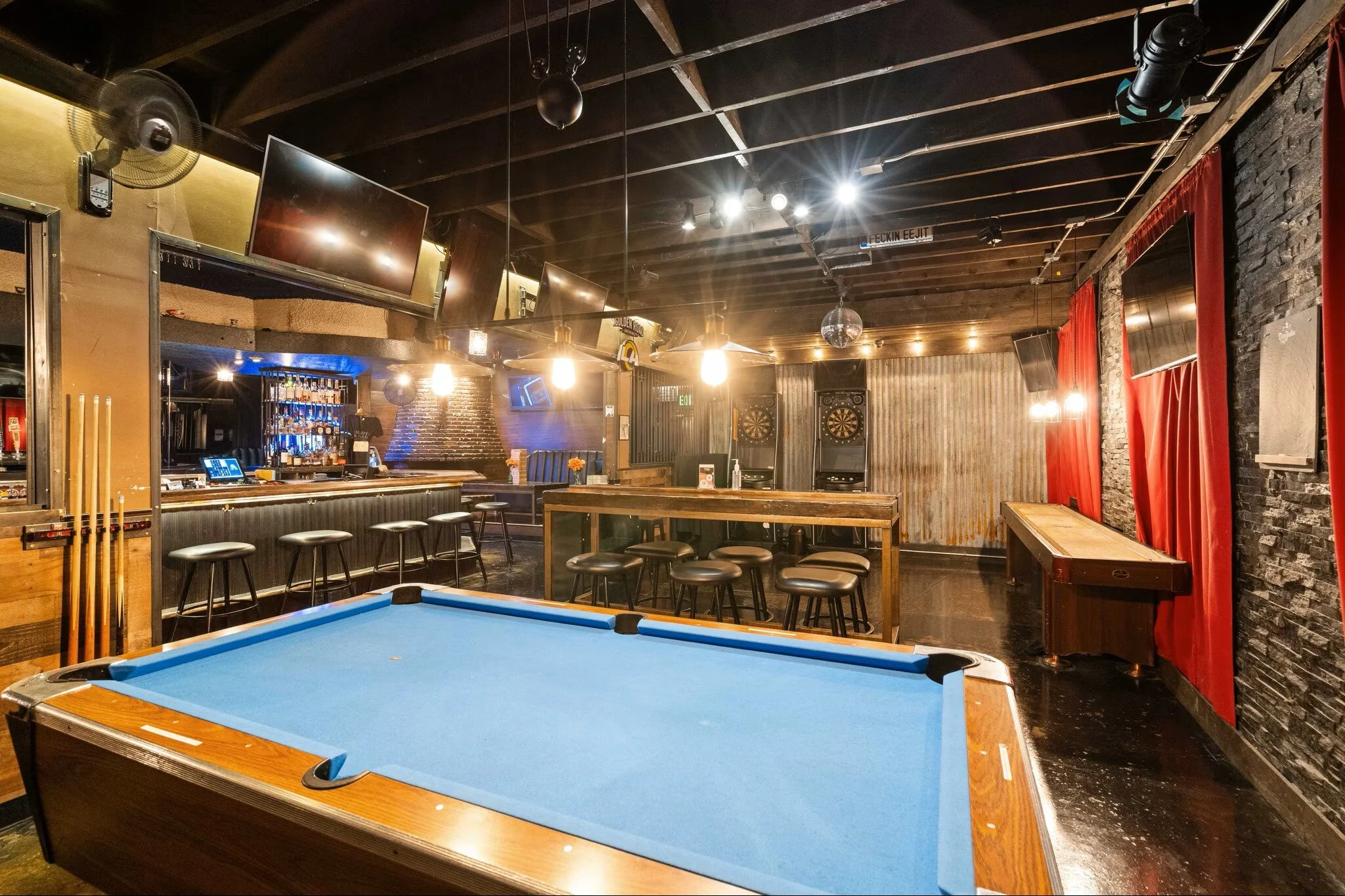 Interior of a bar with a pool table in the foreground, multiple flatscreen TVs mounted on the ceiling, dartboards on the back wall, bar counter with barstools, and red curtains on the right side.