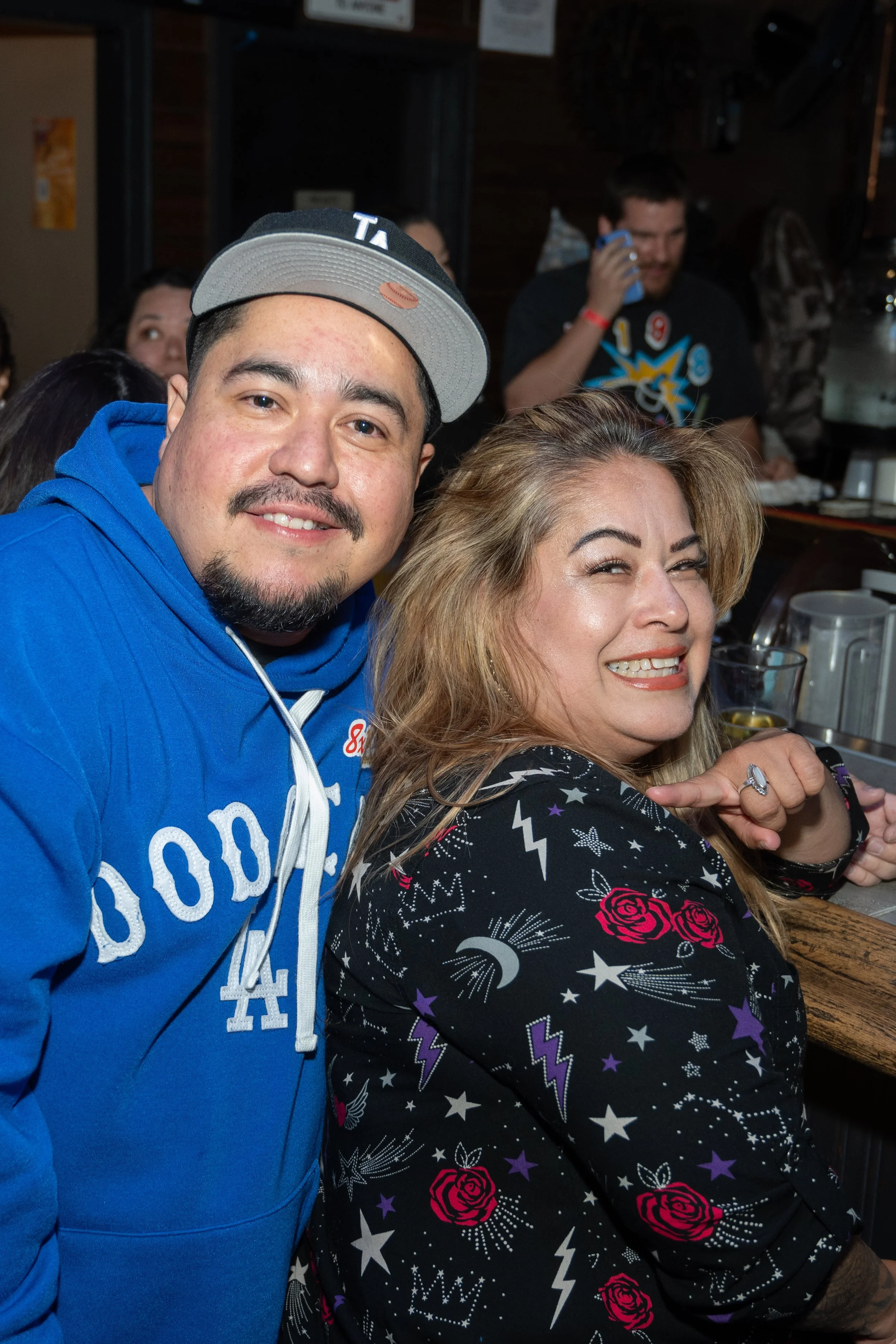 Two people smiling at a bar, one wearing a blue hoodie and cap, the other in a patterned jacket with roses and stars.