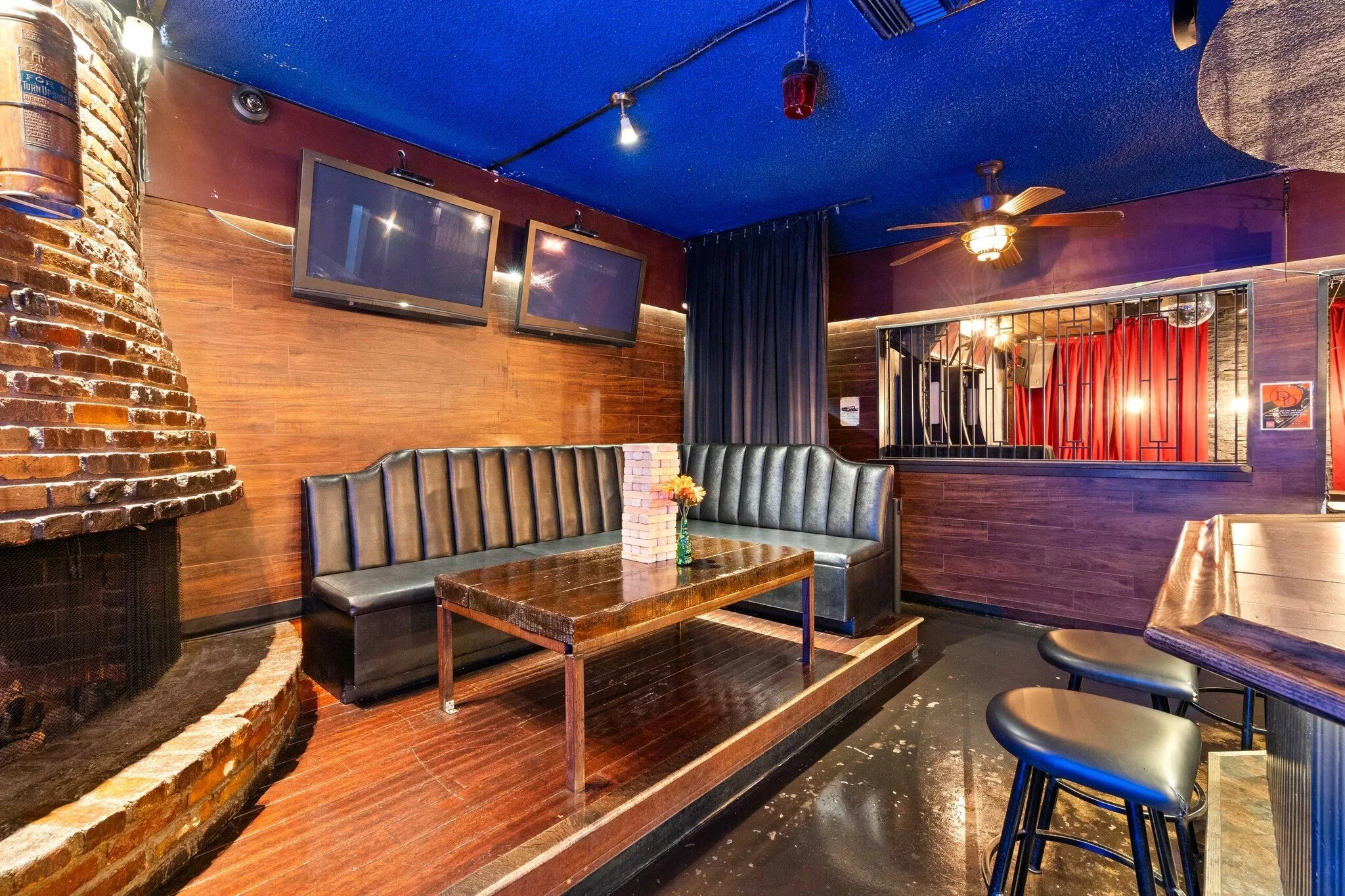 Cozy lounge area with a brick fireplace, a black leather booth, a wooden table with a small flower arrangement, and wall-mounted TVs in a dimly lit bar or restaurant.