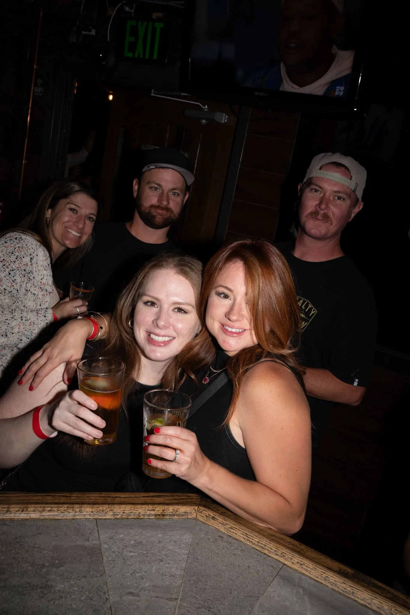 Group of five friends at a bar, smiling and holding drinks, with a TV screen and an exit sign in the background.