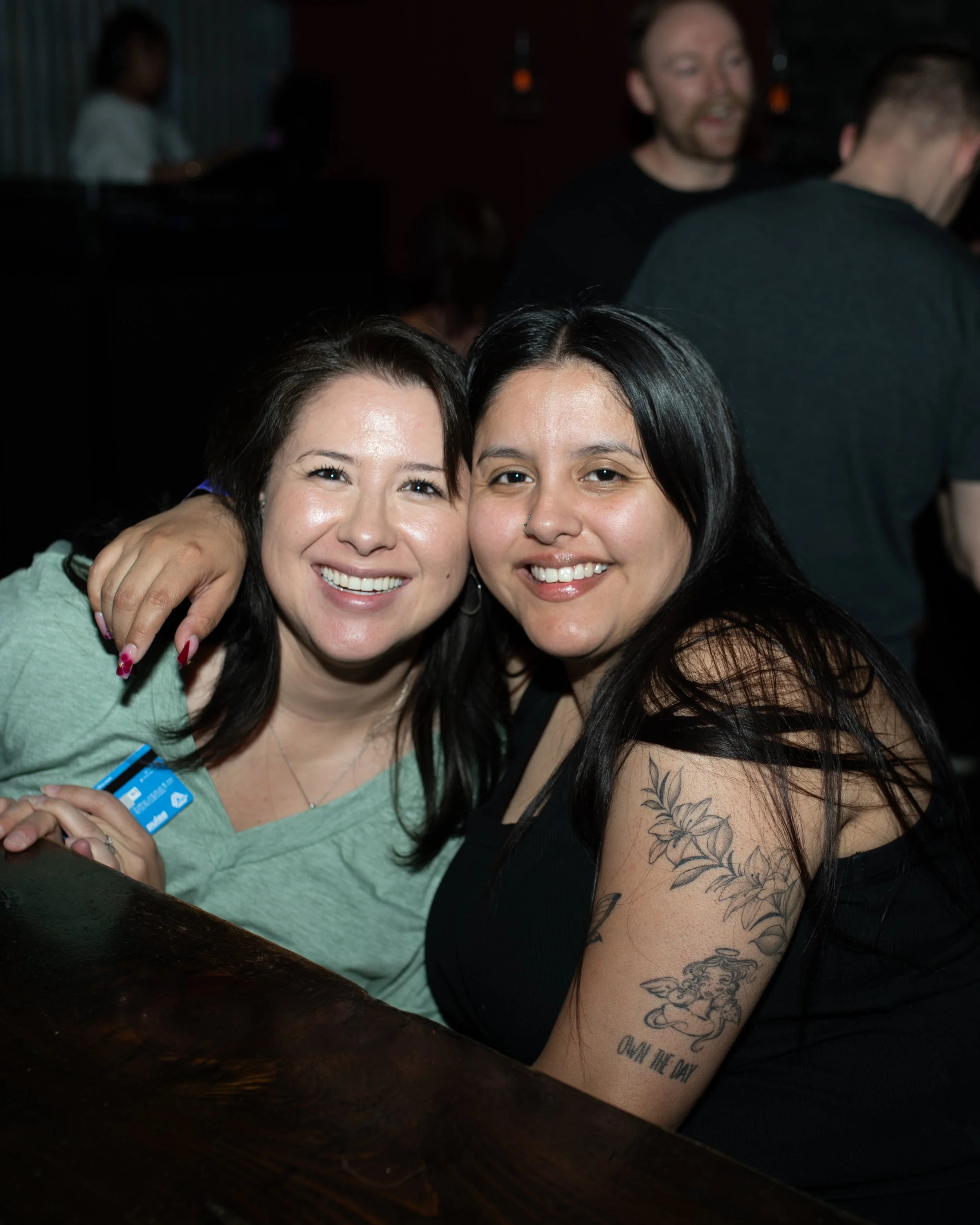 Two women smiling and hugging at a bar or nightclub, with blurred people in the background. One woman has dark hair and tattoos on her arm, wearing a black top. The other woman has dark hair, wearing a light green shirt and a name badge.