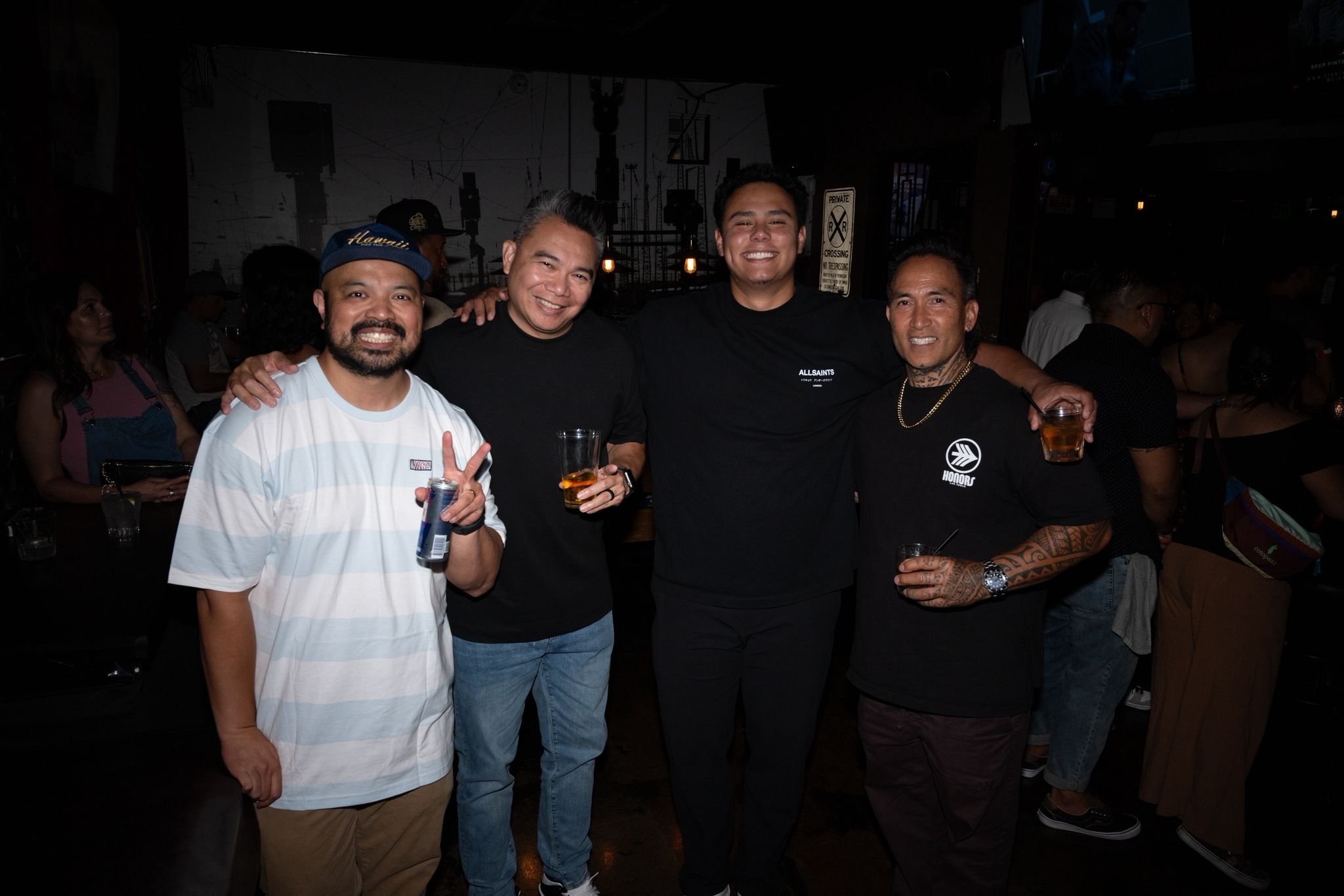 Group of five men at a social gathering, smiling and holding drinks, in a dimly lit indoor venue.