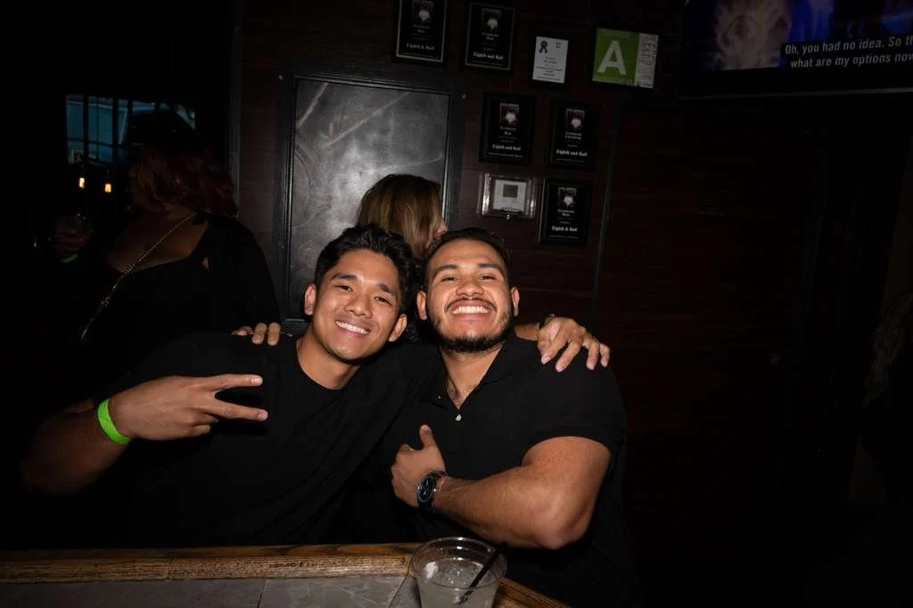 Two men smiling and sitting together in a bar or nightclub, with other people in the background.