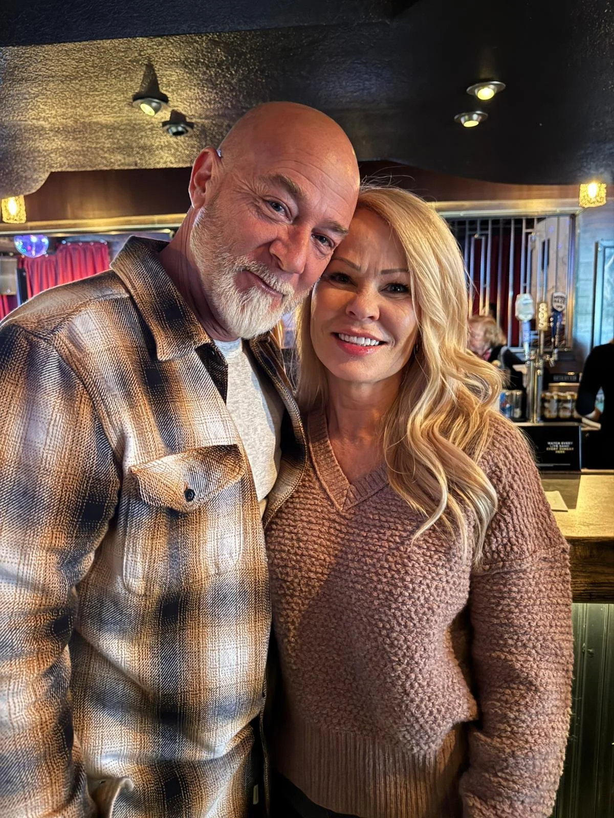 A man with a white beard and a woman with blonde hair smiling, standing close together inside a dimly lit bar or pub.