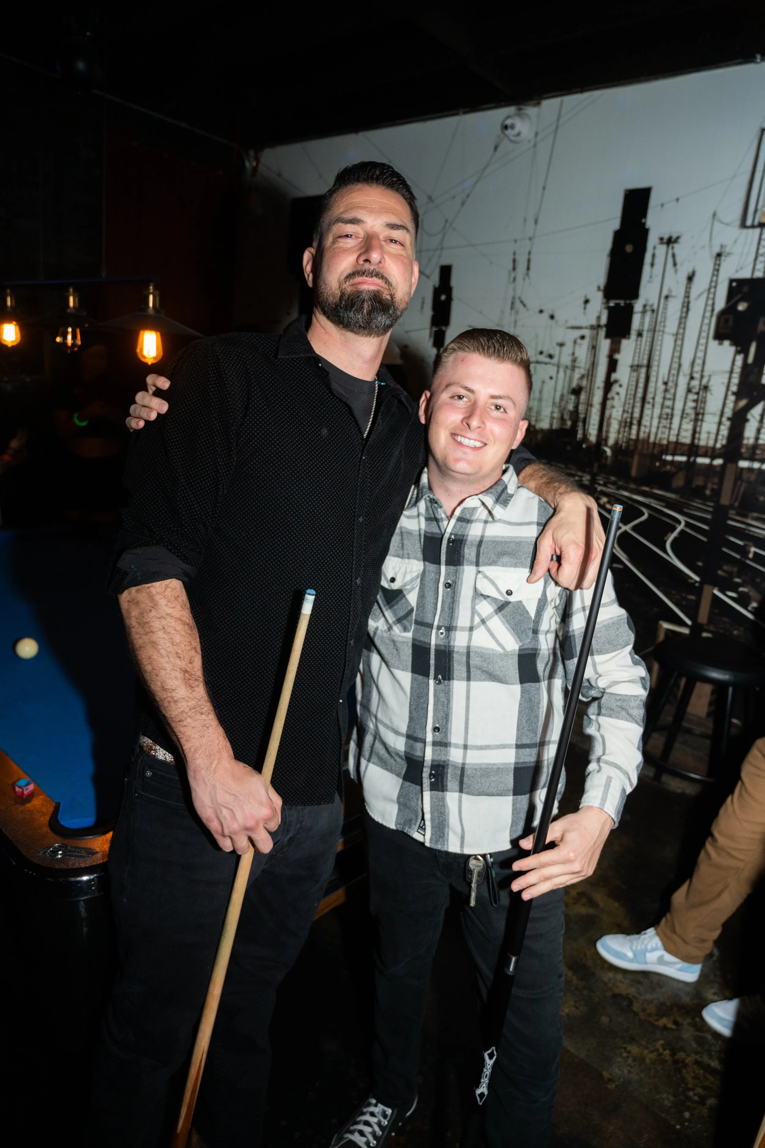 Two men posing with pool cues at a pool table, smiling, with a mural of train tracks in the background.