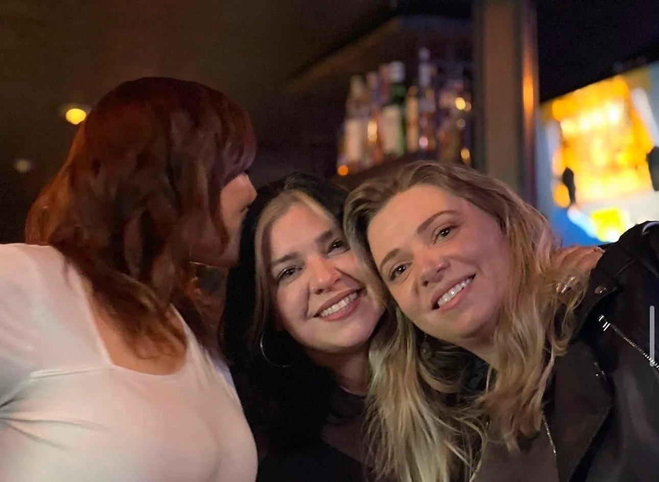 Three women smiling and posing together in a dimly lit bar environment with a shelf of bottles in the background.