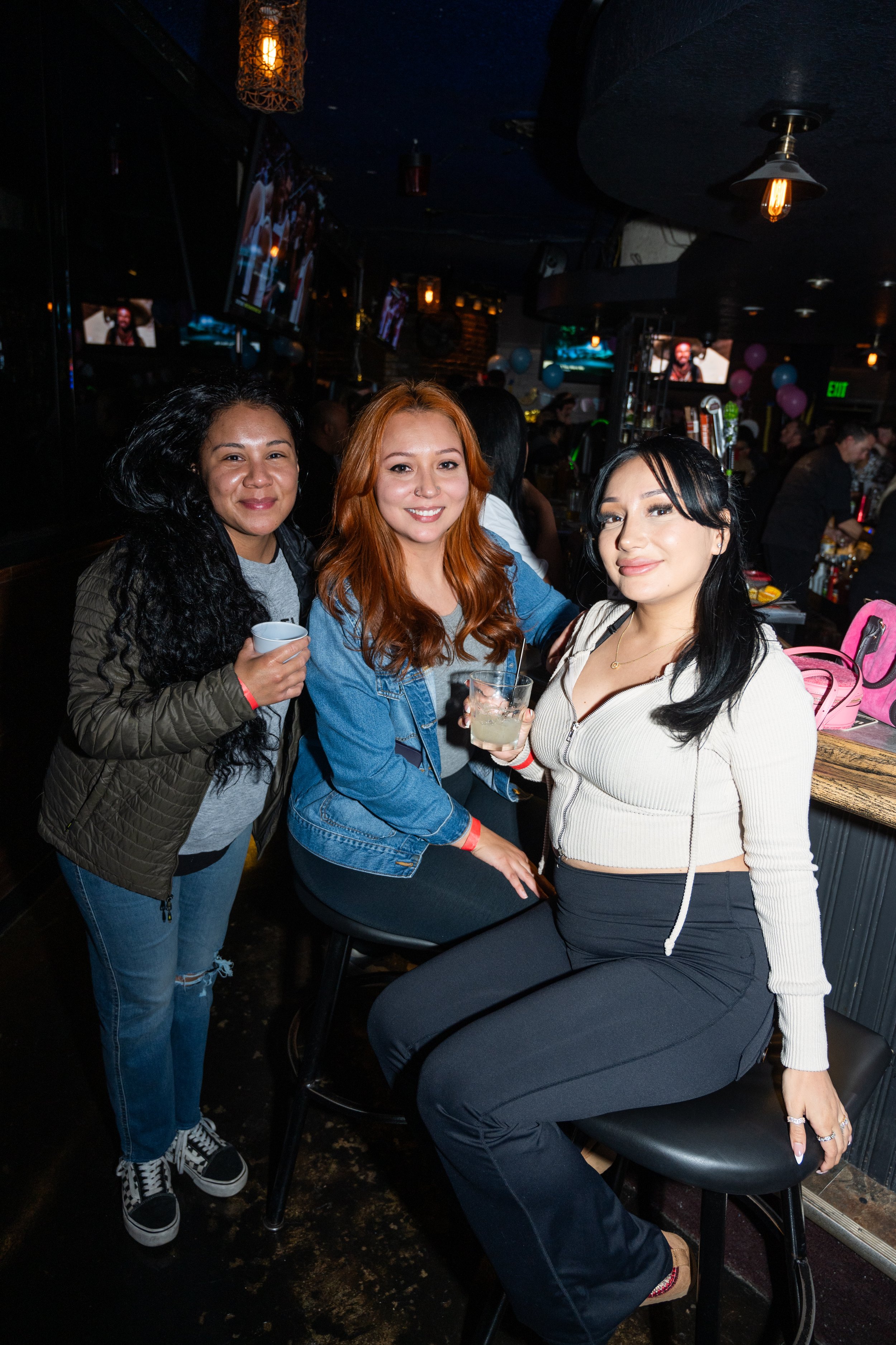 Three women smiling and posing together in a bar setting, holding drinks. The background features dim lighting, decorations, and other patrons in a social atmosphere.