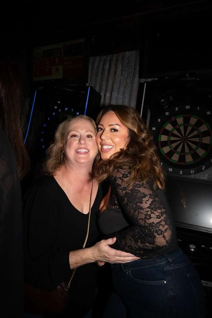 Two women smiling while posing close together in front of a dartboard in a dimly lit bar or pub.