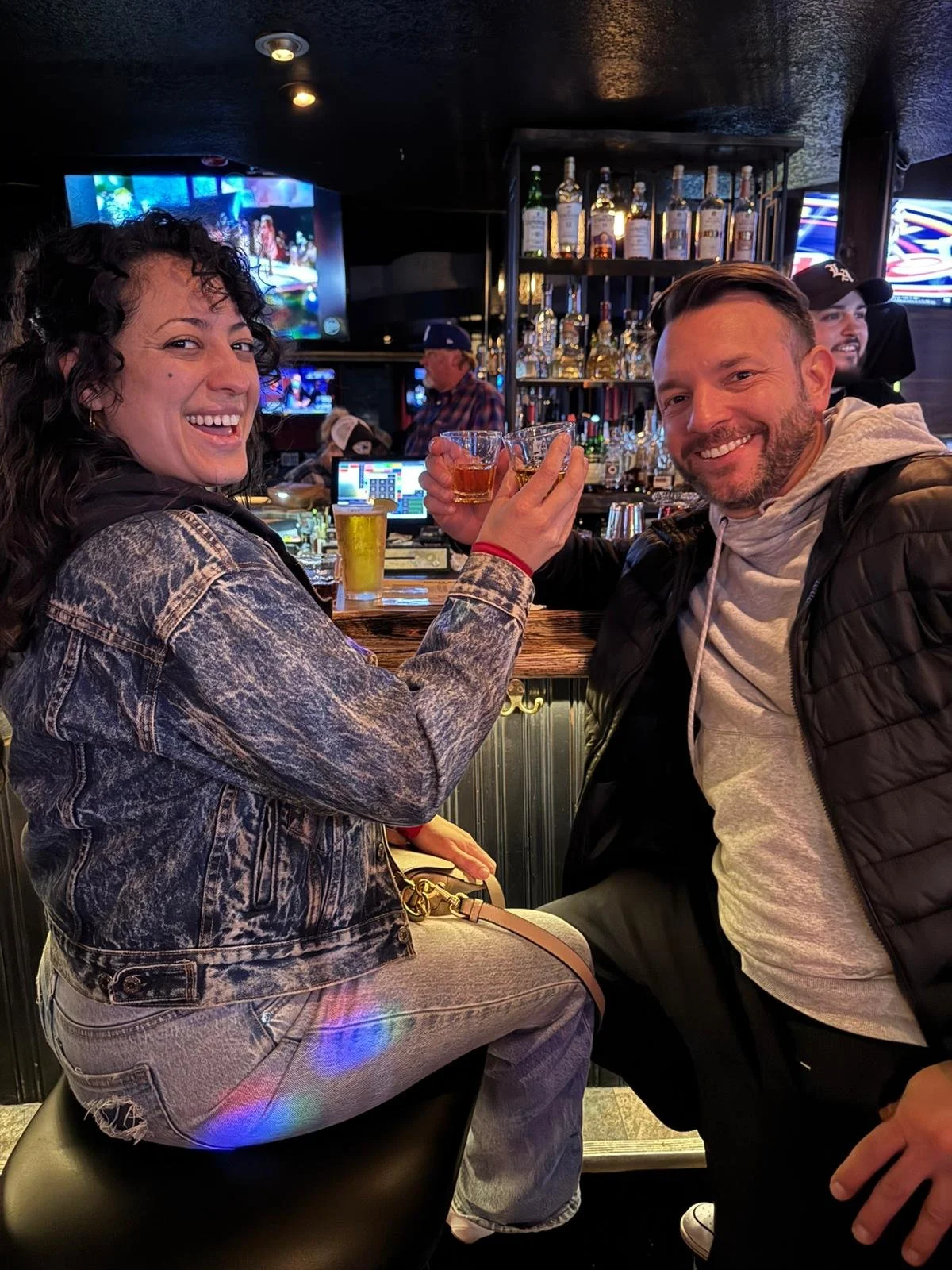 Two smiling people in a bar toasting with drinks, woman on the left with curly dark hair wearing a denim jacket and jeans, man on the right with short hair and beard wearing a gray hoodie and black jacket, bar with bottles and TVs in the background.