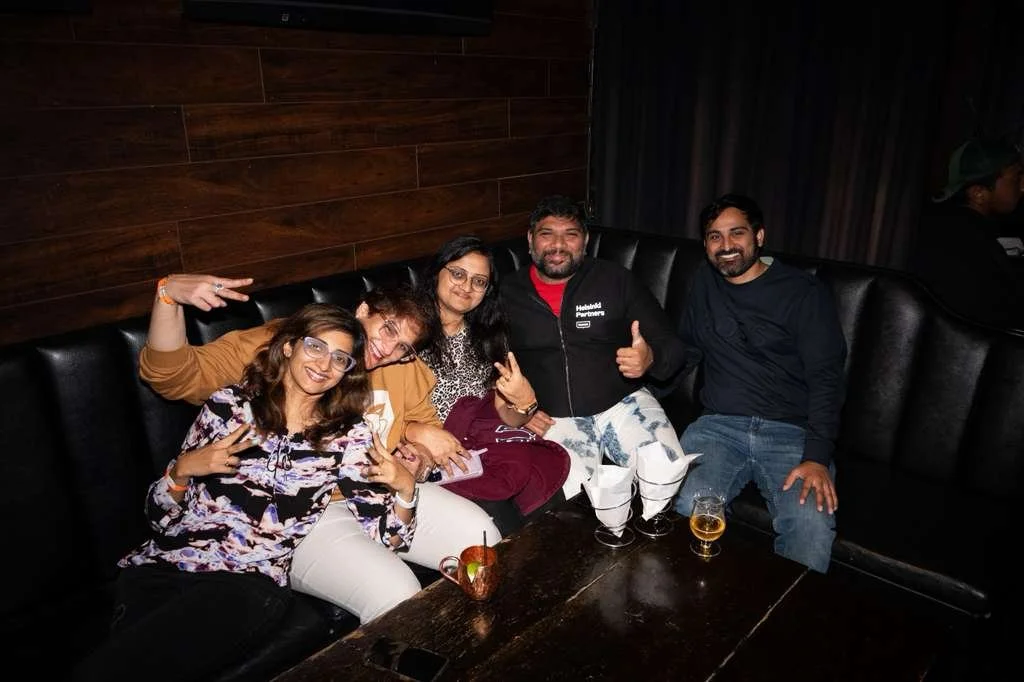 Group of five friends sitting on a black leather booth at a restaurant, smiling and making gestures. Two women are on the left, one wearing glasses and a floral blouse, the other in a tan jacket. The man in the middle is wearing a black jacket with a