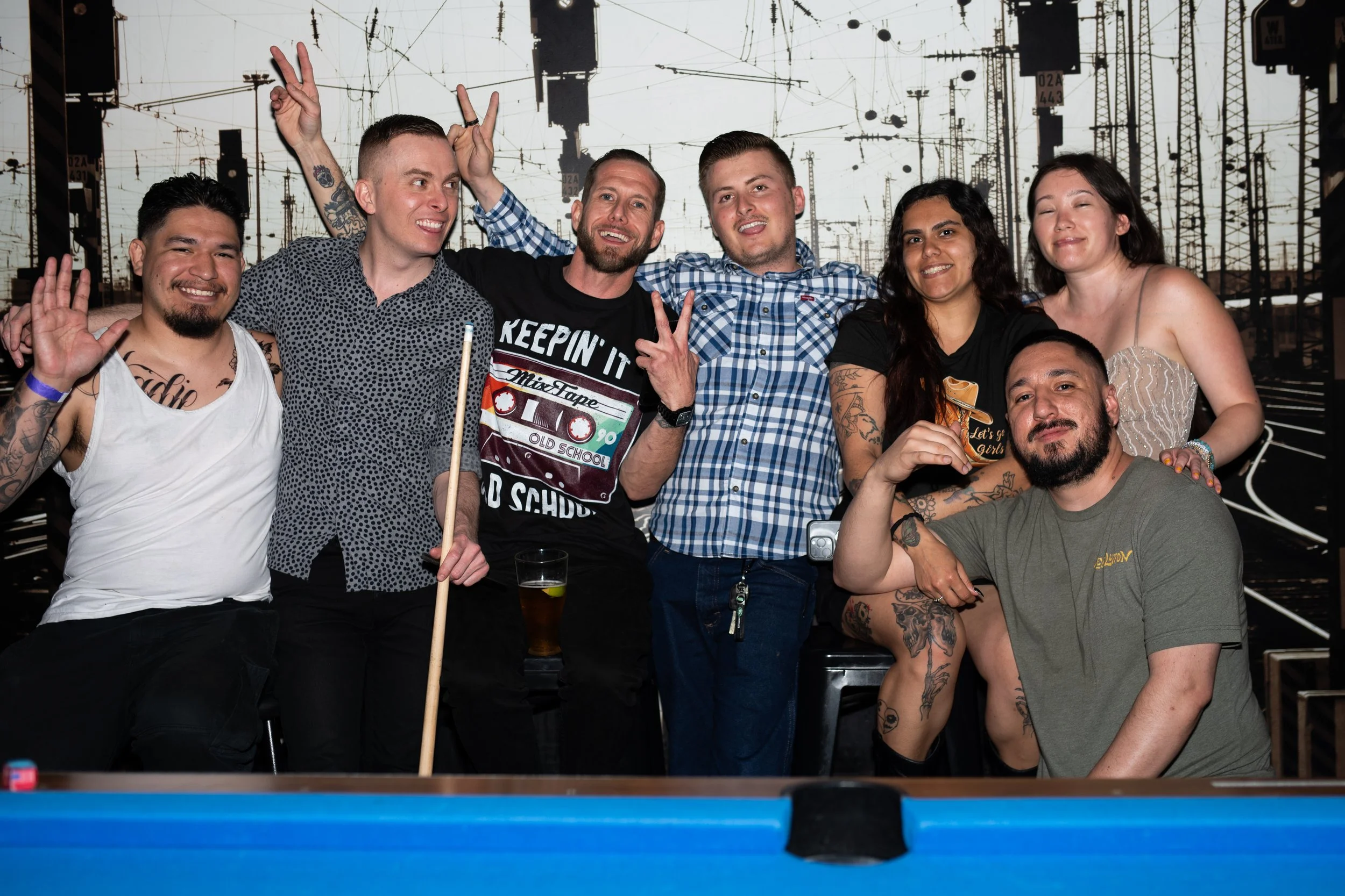 A group of eight diverse young adults celebrating around a pool table with a cityscape background, some making peace signs and smiling.