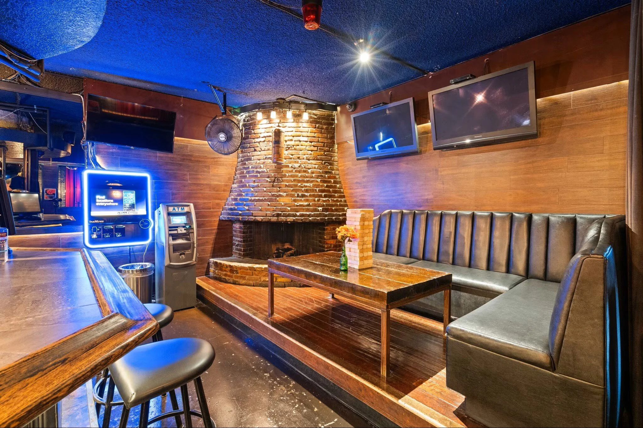 Cozy bar with brick fireplace, wooden walls, multiple television screens, neon sign, ATM, and bar seating area with stools