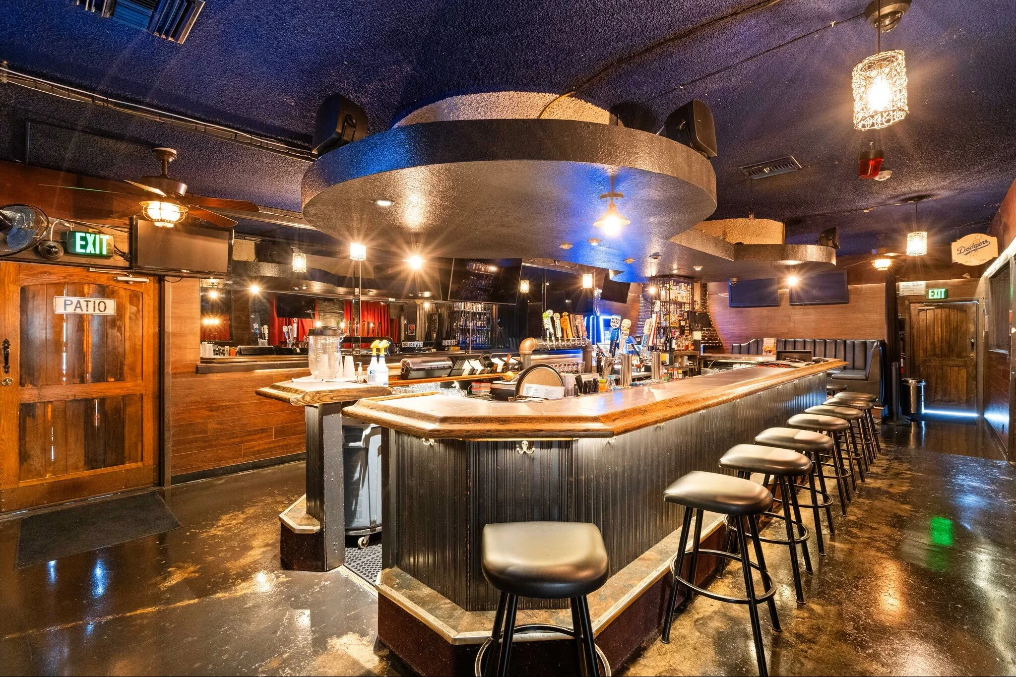 Empty bar with a curved wooden counter, black barstools, multiple tap handles, and warm ambient lighting in a dimly lit pub or restaurant.