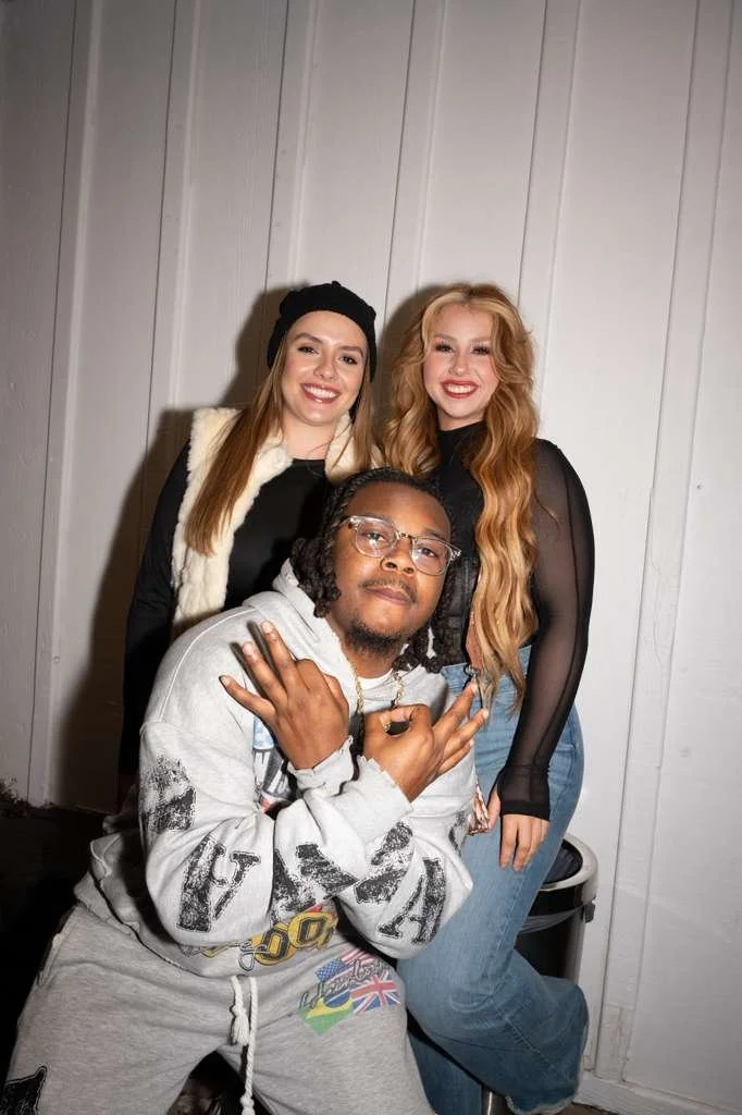 Three young adults posing for a photo indoors against a white paneled wall, smiling and making hand gestures.