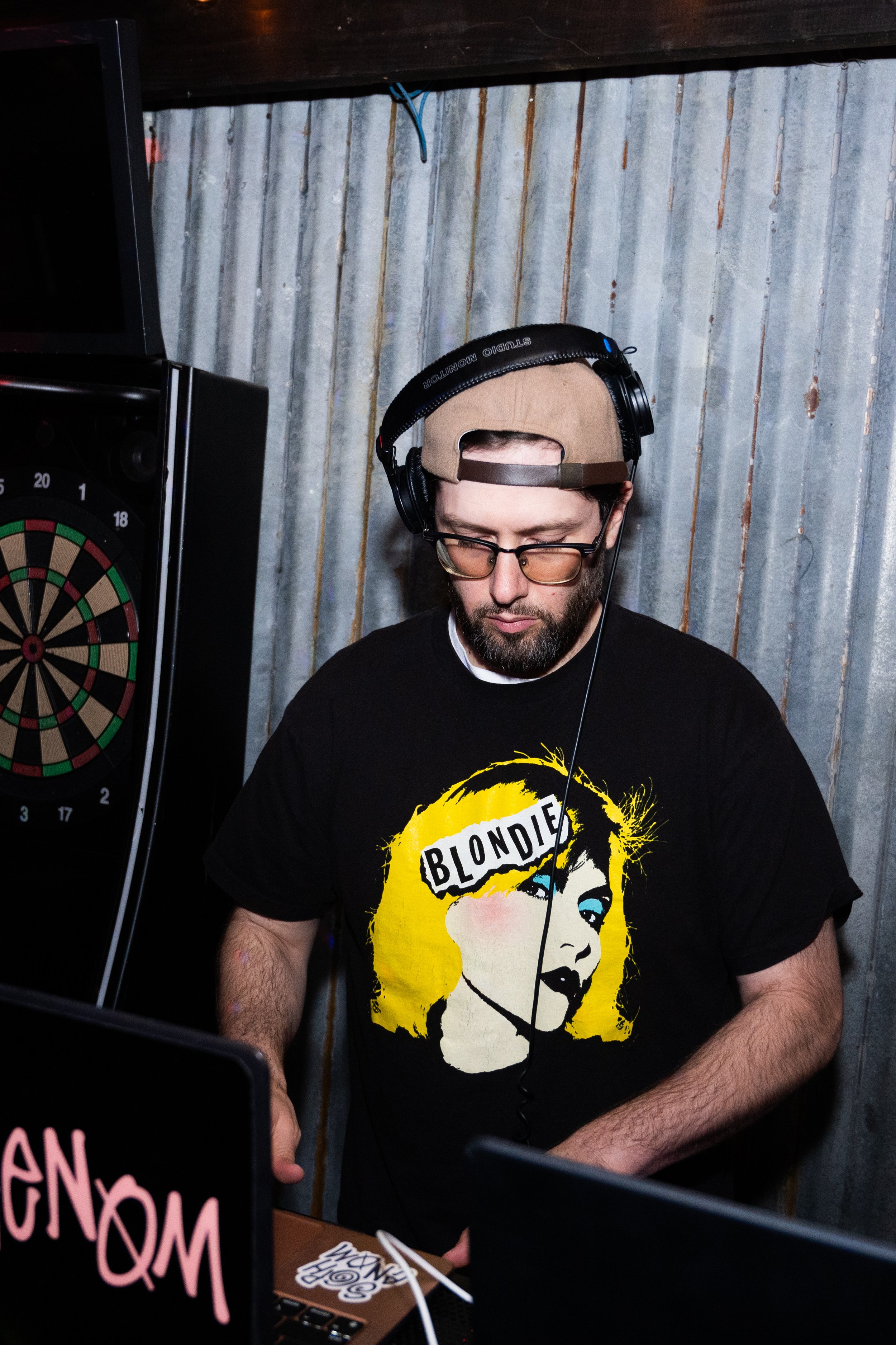DJ wearing headphones and a Blondie t-shirt, standing near a dartboard and DJ equipment in a dimly lit room with corrugated metal walls.