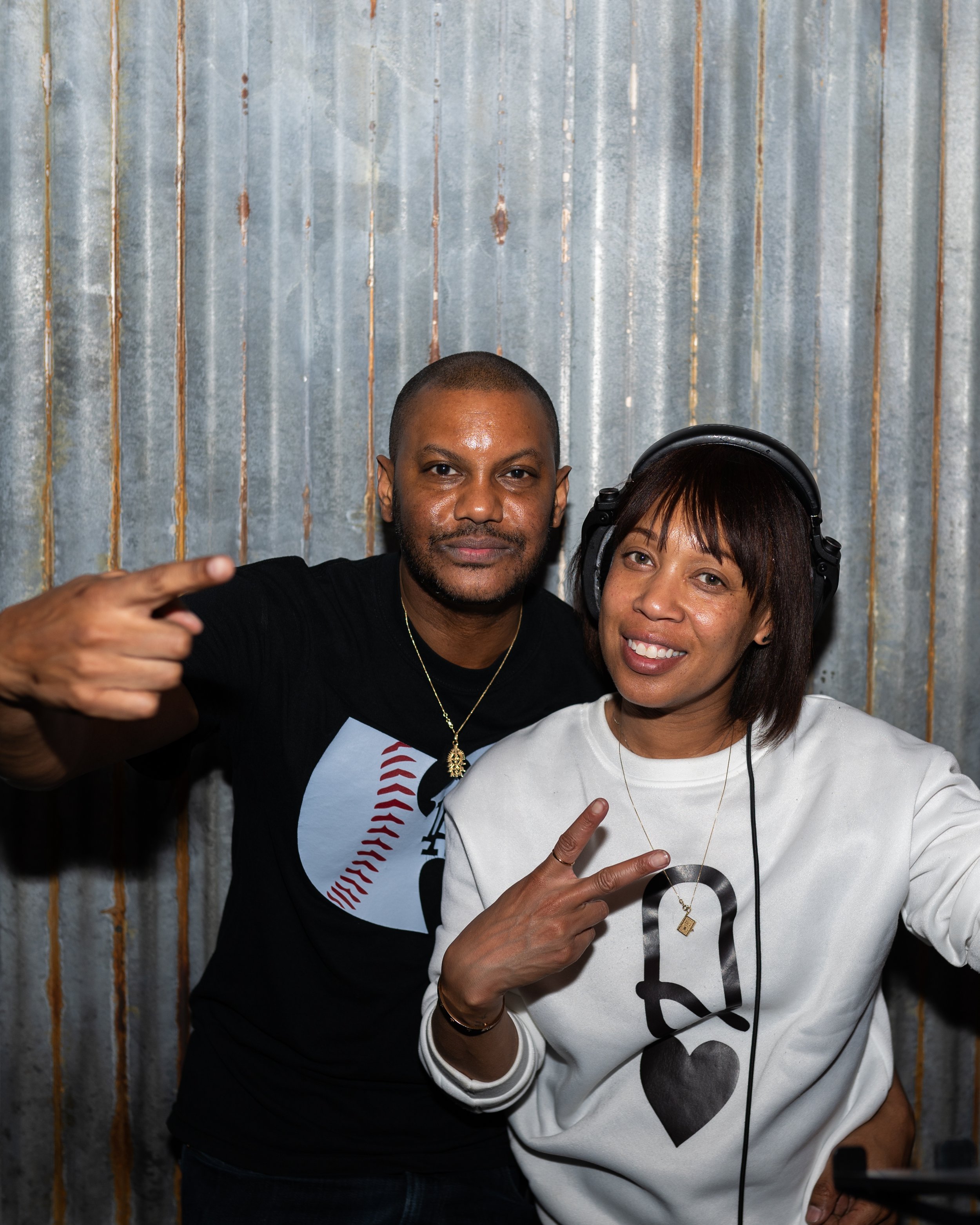 Two people, a man and a woman, are posing together in front of a corrugated metal wall. The man is pointing to himself with his left hand, and the woman is holding up a peace sign with her right hand. The woman is wearing large headphones, and both a