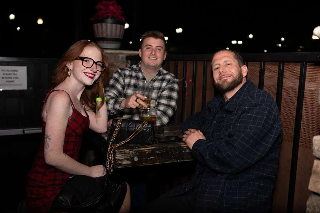 Three people sitting together at an outdoor table at night, smiling and holding drinks, with decorative lights and a potted plant in the background.