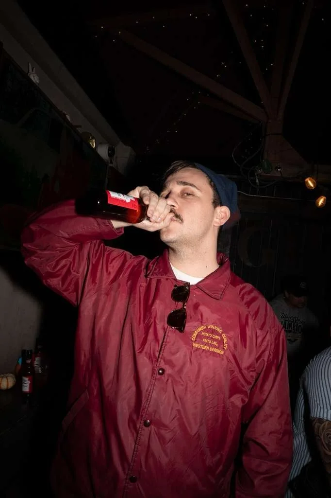 A man in a red jacket and a blue cap is drinking from a brown bottle, standing indoors at a party or social gathering.