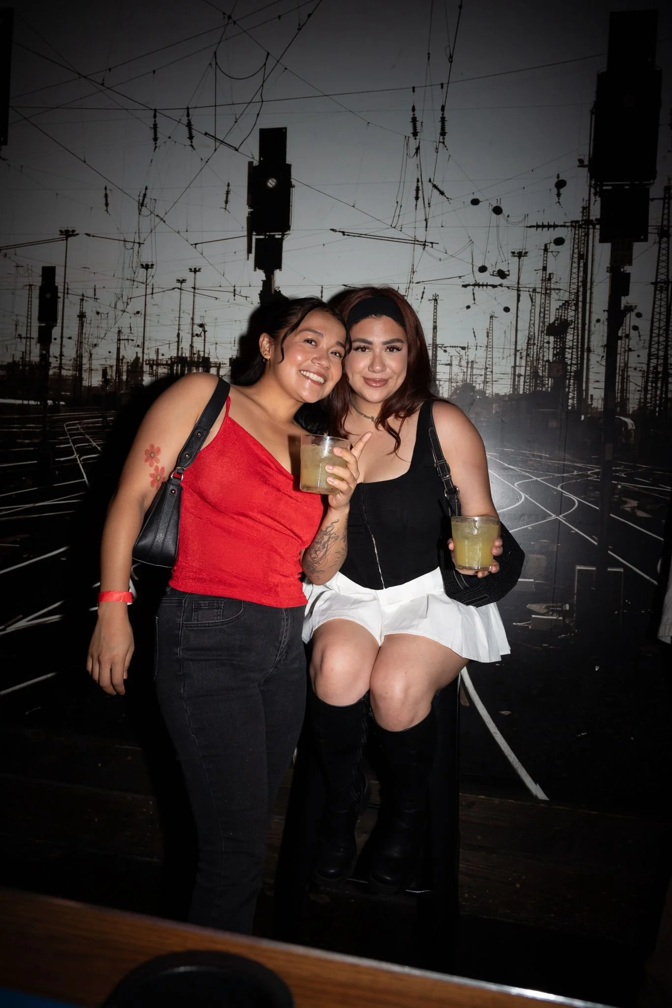 Two women smiling and holding drinks in a dark environment with a backdrop of railway tracks and overhead wires.