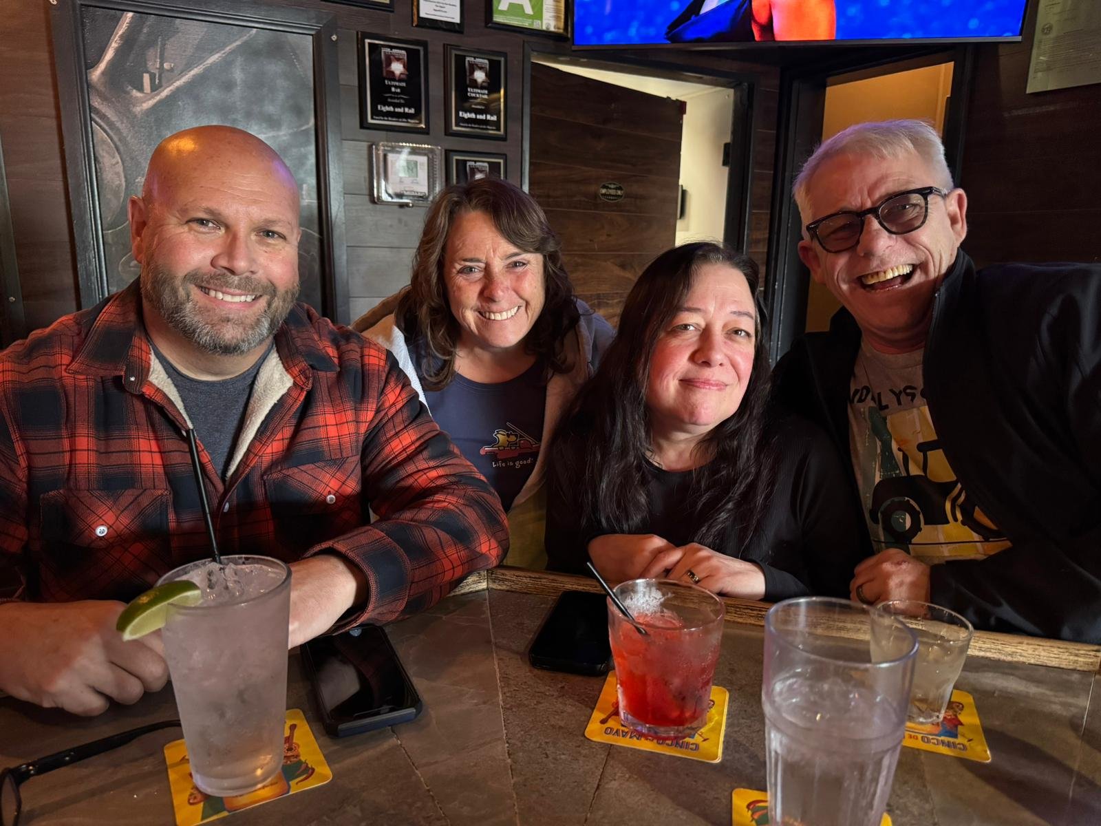 Four people sitting at a table in a restaurant, smiling for the camera. There are drinks on the table, and a dark wooden wall with framed pictures in the background.