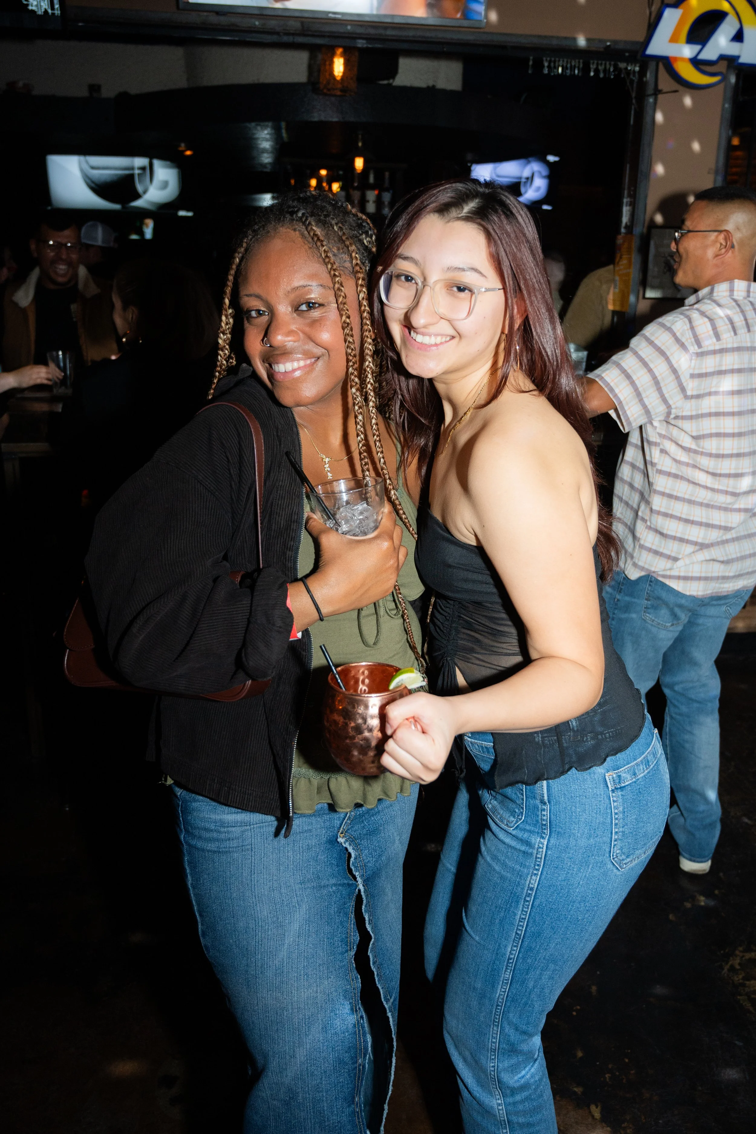 Two women smiling and posing for a photo at a bar, holding drinks. The background shows people socializing. They are wearing casual outfits and appear to be enjoying a night out.